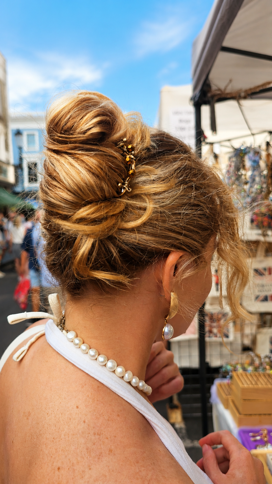 A woman with blonde hair styled in a high, messy bun with a small gold hair accessory, wearing a white halter top and a pearl necklace, photographed from behind in an outdoor market setting.