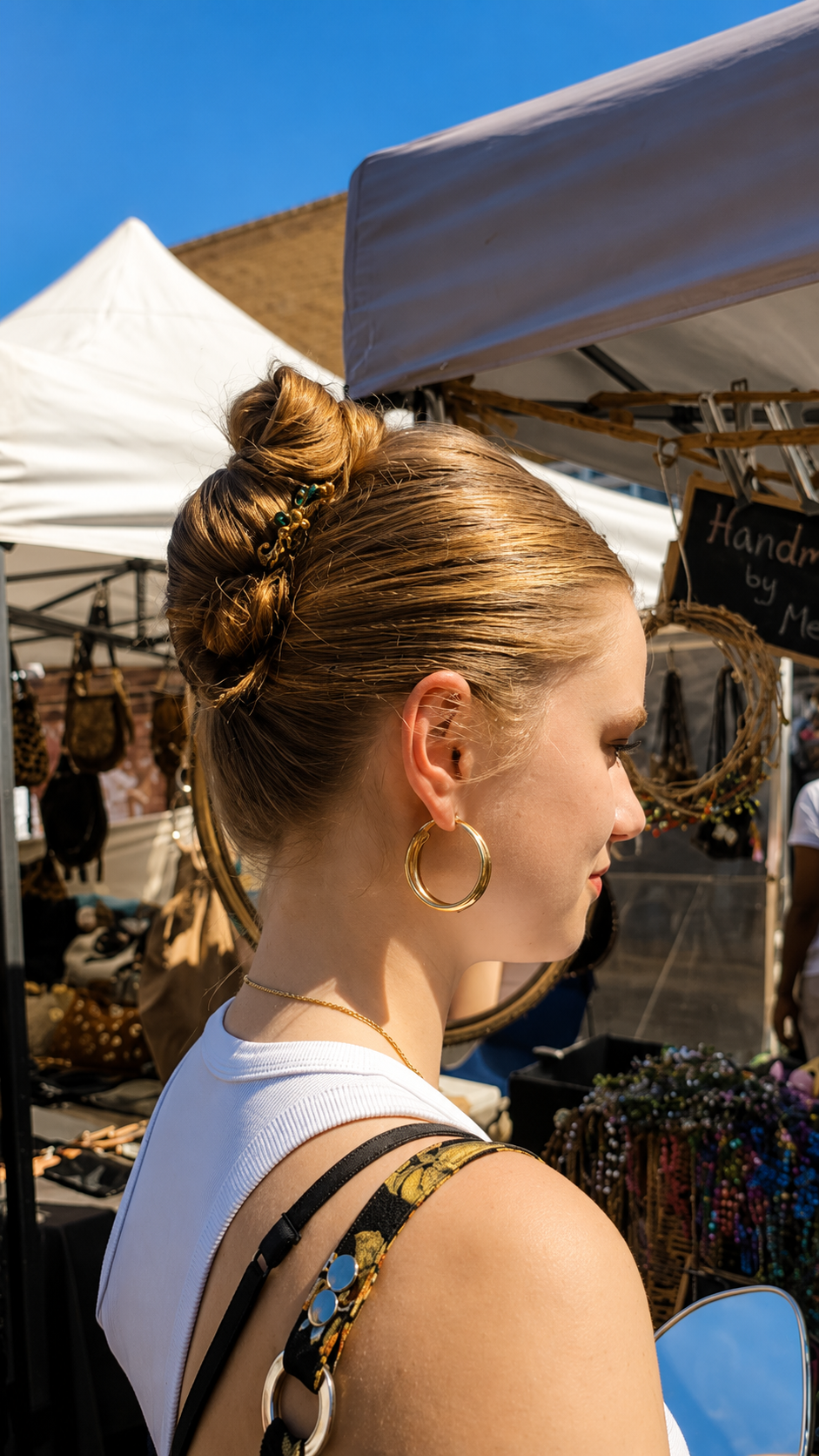 A woman with blonde hair in a messy bun is shown in profile at an outdoor market, wearing a gold hoop earring and a white top. Market stalls and tents are visible in the background under a blue sky.