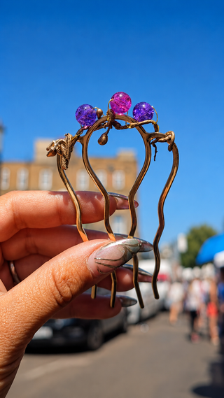 A hand with silver glittery nails holds a gold metal hair comb decorated with three colorful gemstones in purple, pink, and blue, set against a blurred outdoor street background with a blue sky.