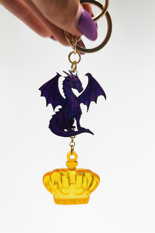 A keychain featuring a detailed purple dragon charm paired with a glossy golden crown pendant, connected by a gold chain on a white background