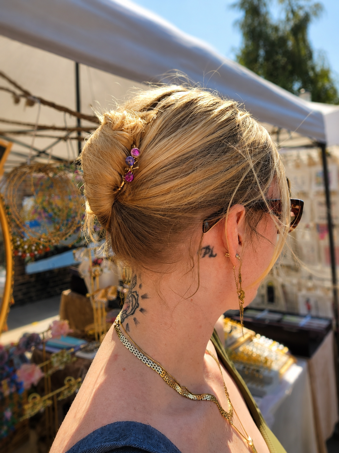 A woman with blonde hair in a bun wearing a small pink floral hair accessory, shown from the side at an outdoor market under a white canopy.